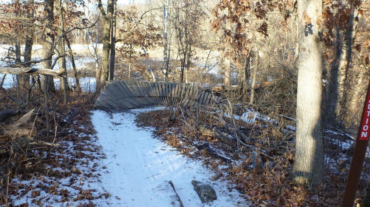 A snowy path surrounded by trees, featuring a damaged wooden barrier bent at an angle. The ground is covered with fallen leaves, and a caution sign is visible on the right side of the image, indicating potential hazards in the area. Hillside Park mountain bike trail.
