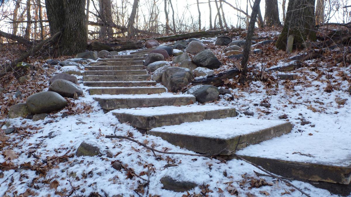 Stone steps lead up a snowy path in a wooded area, surrounded by large rocks and fallen leaves. The scene captures a serene winter atmosphere with bare trees in the background. Hillside Park mountain bike trail.