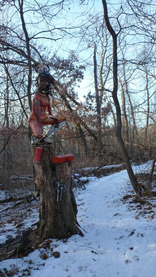 A wooden sculpture of a seated figure with intricate details, dressed in orange and gray attire, perched atop a tree stump in a forested area. The surrounding landscape is wintery, with snow covering the ground and leafless trees in the background. A winding path can be seen leading through the woods. Hillside Park mountain bike trail.