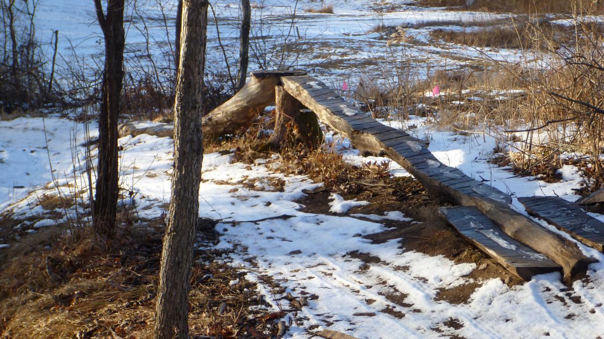 A wooden bridge made from logs and planks spans a small area, surrounded by a mix of snow and bare ground. There are trees and brush nearby, and a few pink flags are visible in the background, indicating nearby markers or trails. The scene captures a winter landscape with a natural, rustic feel. Hillside Park mountain bike trail.