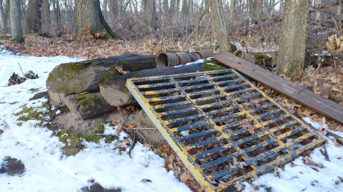A discarded metal grate lies on the ground near a large stone in a snowy, wooded area. Surrounding the grate are patches of brown leaves and fallen tree branches. In the background, bare trees rise up, indicating a winter landscape. Hillside Park mountain bike trail.