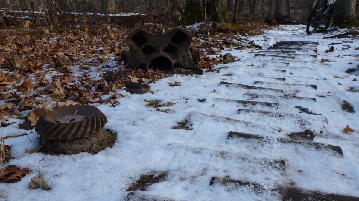 A snowy pathway lined with fallen leaves, featuring rusty mechanical gear parts and an old metal object on the side. In the background, trees are visible, and a mountain bike leans against a tree. Hillside Park mountain bike trail.