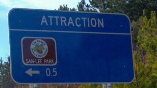 Sign indicating an attraction with the text "ATTRACTION" at the top, a logo of Sam-Lee Park, and an arrow pointing left with a distance of 0.5 miles. San-lee Park mountain bike trail.
