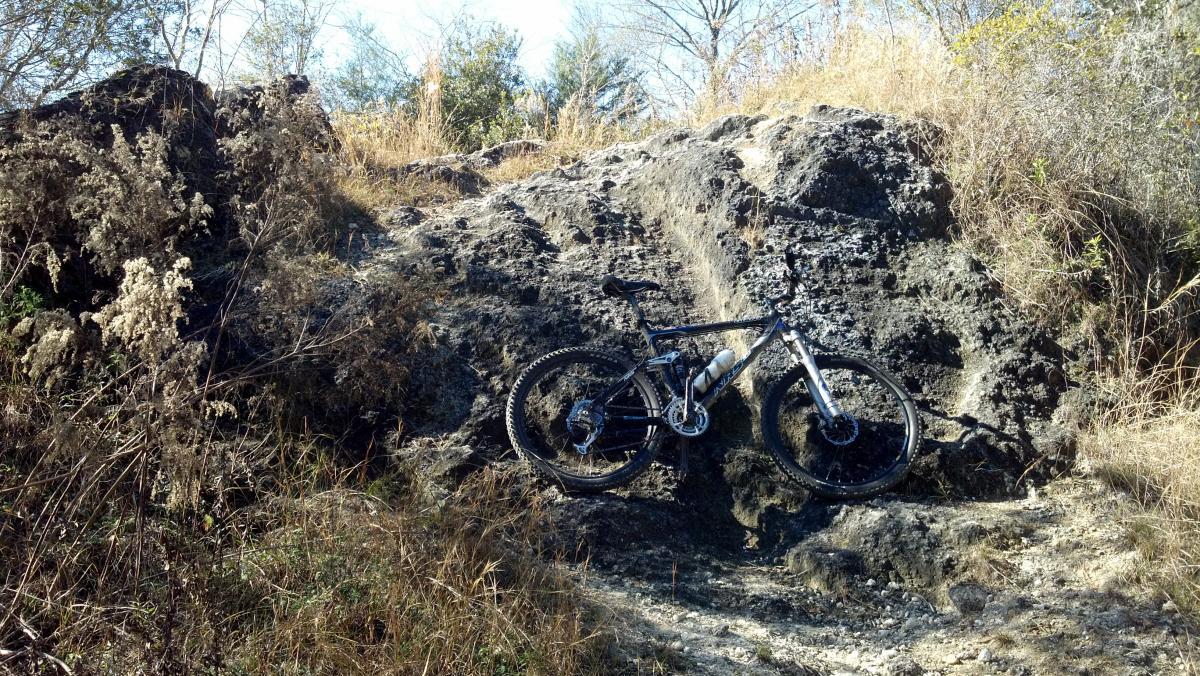 A mountain bike leaning against a rocky outcrop, surrounded by tall grass and scattered vegetation in a natural setting. The scene captures a sunny day with clear skies, suggesting an outdoor adventure location. Santos mountain bike trail.