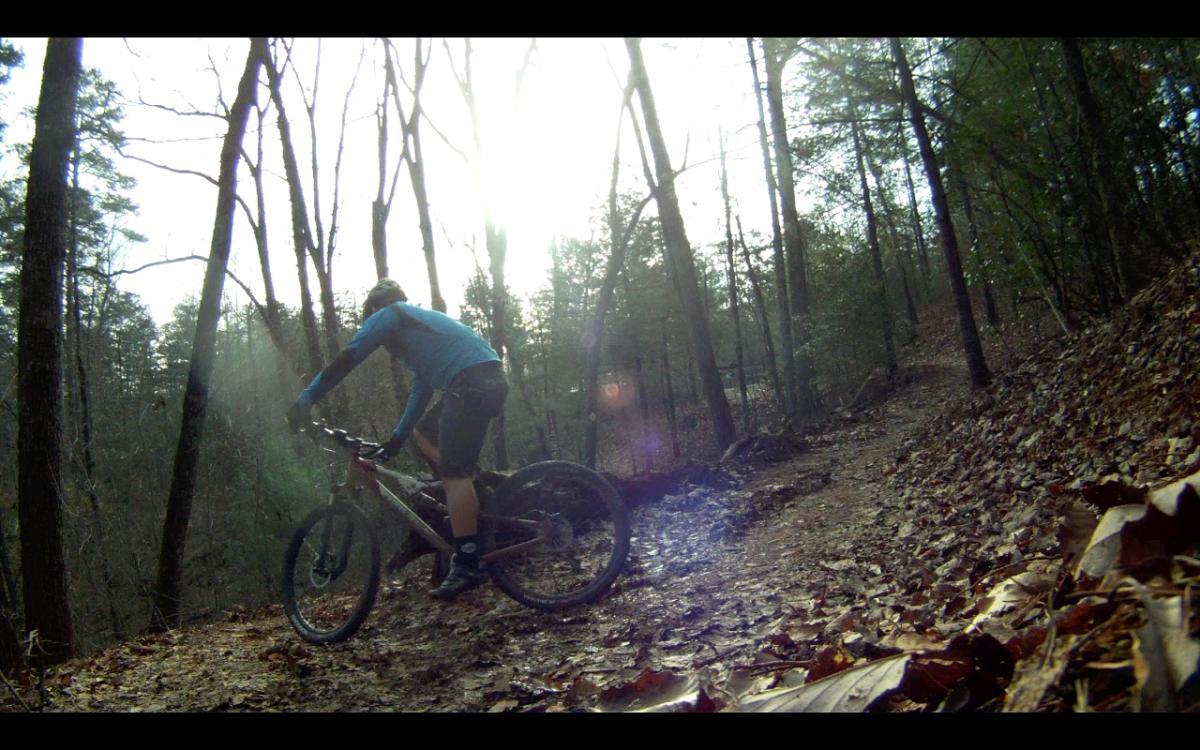A mountain biker navigates a muddy trail surrounded by trees, with sunlight filtering through the branches above. The rider is dressed in a blue long-sleeve shirt and is focused on the path ahead. Fallen leaves cover the ground, adding to the autumnal scenery. Jake Mountain Trails mountain bike trail.