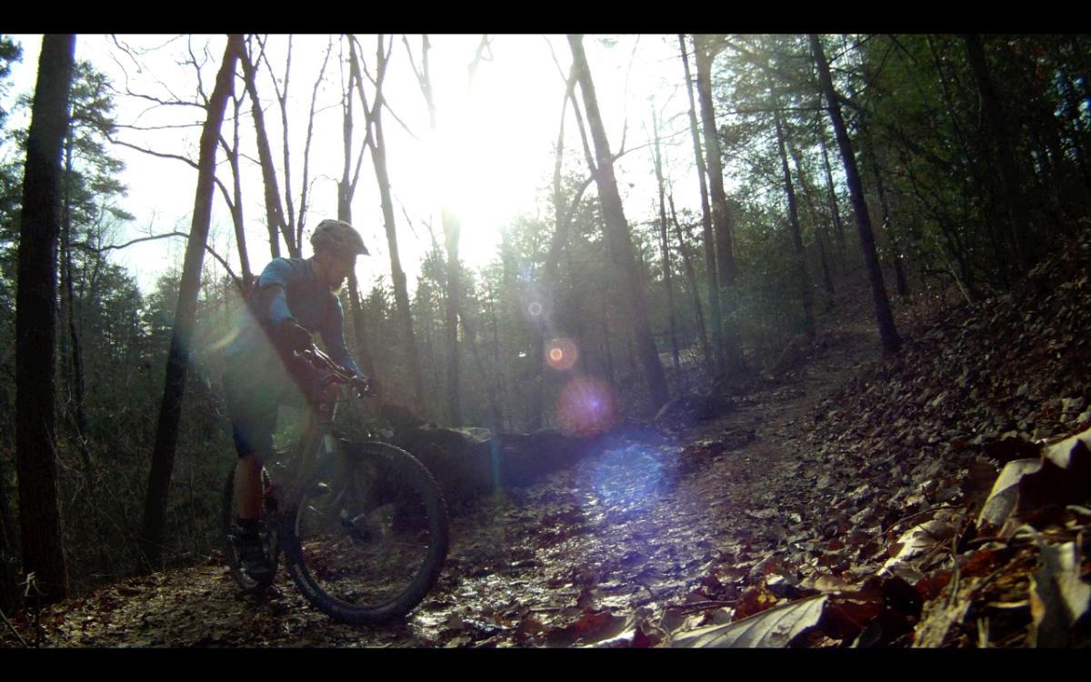 A mountain biker riding along a muddy, leaf-covered trail in a forest, with sunlight filtering through the trees, creating a bright and dynamic atmosphere. Jake Mountain Trails mountain bike trail.