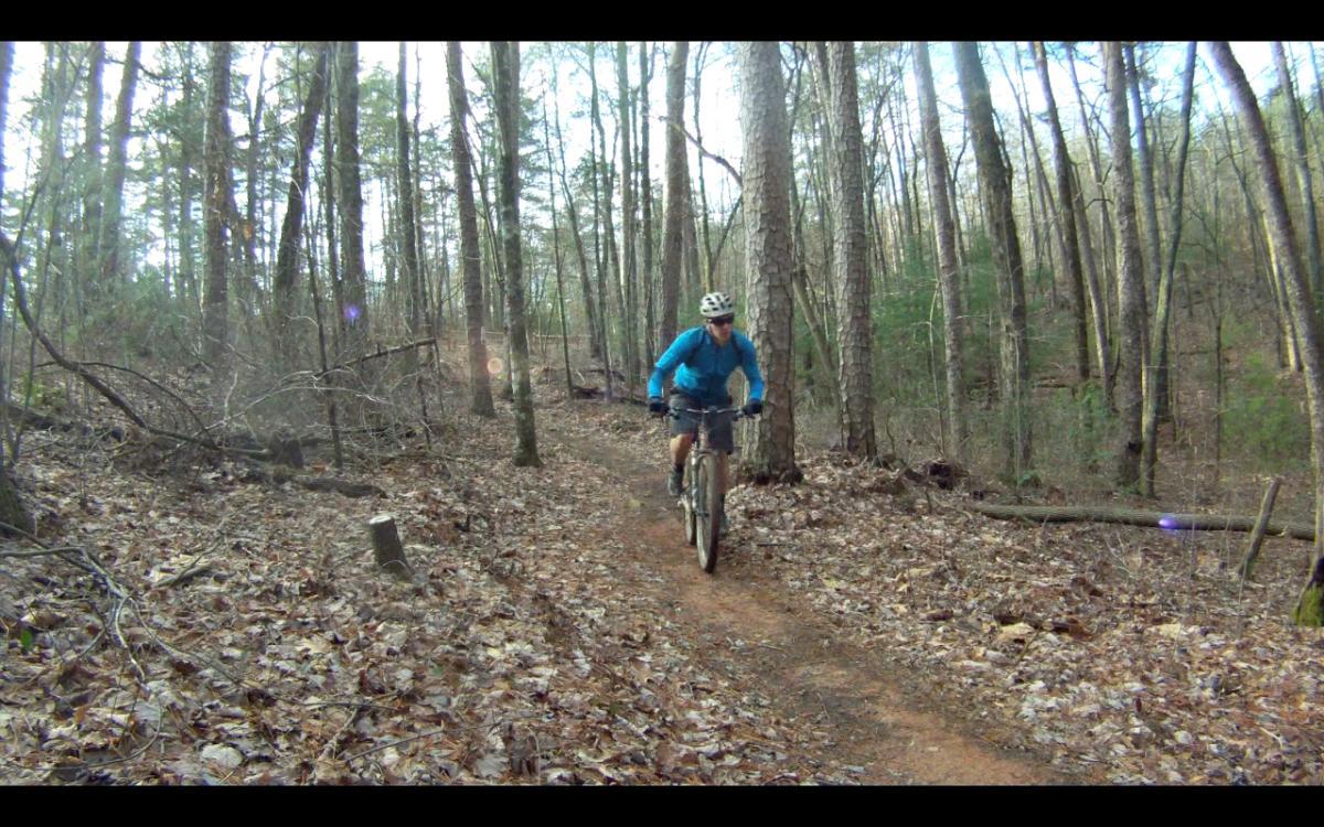 A mountain biker riding along a narrow, dirt trail surrounded by trees in a forested area. The ground is covered with fallen leaves, and the cyclist is wearing a blue jacket and a helmet. The scene captures a natural outdoor setting with a mix of sunlight filtering through the trees. Jake Mountain Trails mountain bike trail.