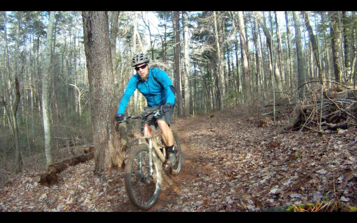 A mountain biker in a blue jacket rides on a dirt trail surrounded by trees in a wooded area. The ground is covered with fallen leaves, and the biker is focused as he navigates the path. Jake Mountain Trails mountain bike trail.