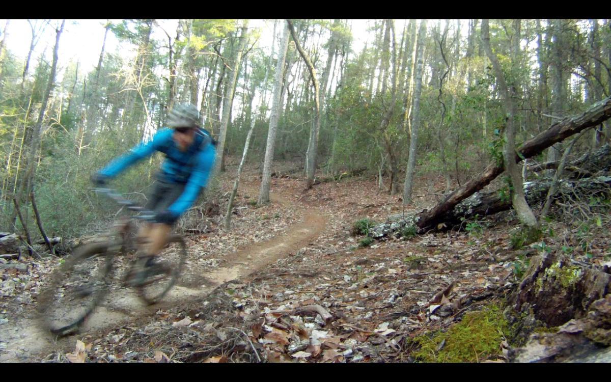 A blurred image of a mountain biker in motion on a narrow dirt trail, surrounded by a wooded area with trees and fallen leaves. The biker is wearing a blue jacket and a hat, navigating a curve in the trail. Soft sunlight filters through the trees, highlighting the natural setting. Jake Mountain Trails mountain bike trail.