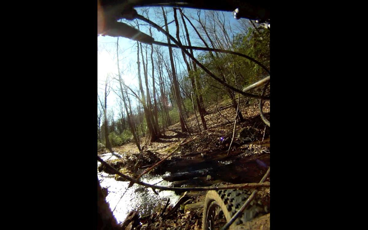 A close-up view of a mountain bike tire navigating over a rocky terrain next to a small stream, with sun shining through trees in a forest setting. Etowah River Trail mountain bike trail.