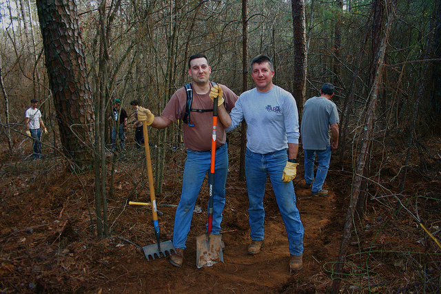 Two men stand smiling in a wooded area, each holding a gardening tool—a shovel and a rake—while others work in the background. The ground is partially cleared, showing a path being created in the forest. The men are dressed casually and wearing gloves, indicating they are engaged in outdoor labor.