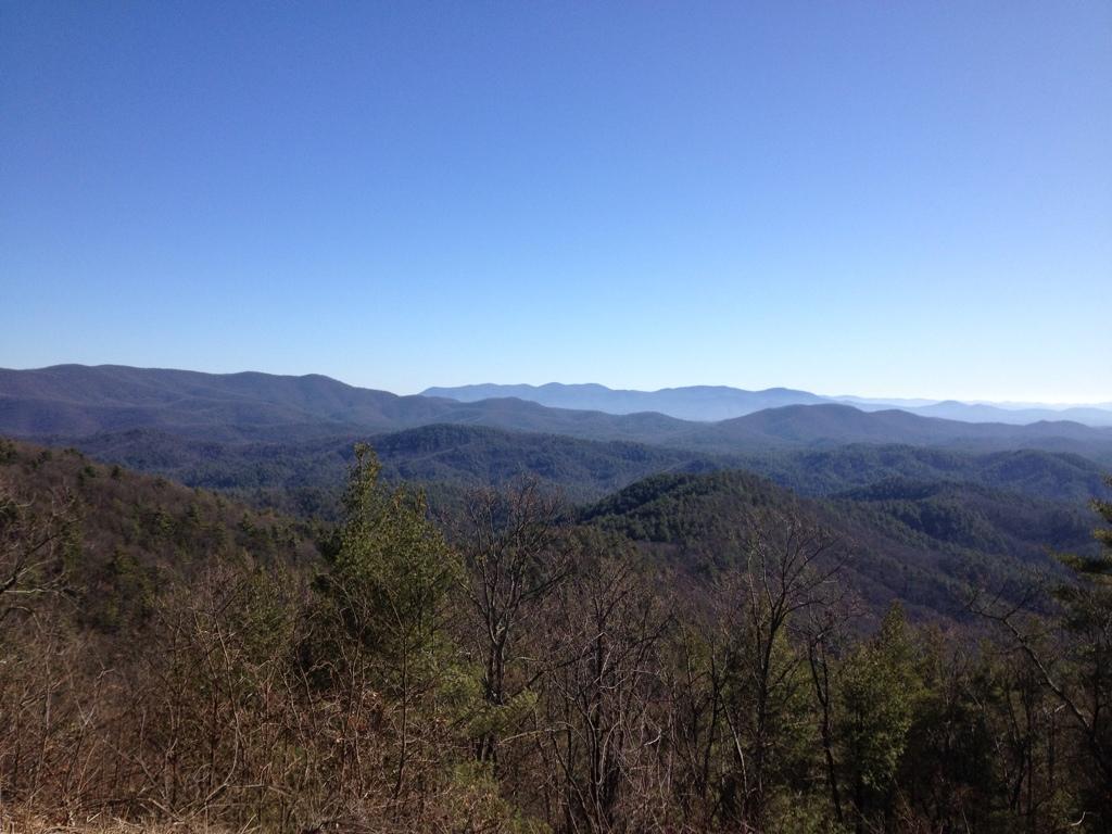 A panoramic view of a mountainous landscape under a clear blue sky, featuring layers of rolling hills and ridges covered in a mix of evergreen and deciduous trees. The scene captures the beauty of nature in a serene and tranquil setting. Bear Creek mountain bike trail.