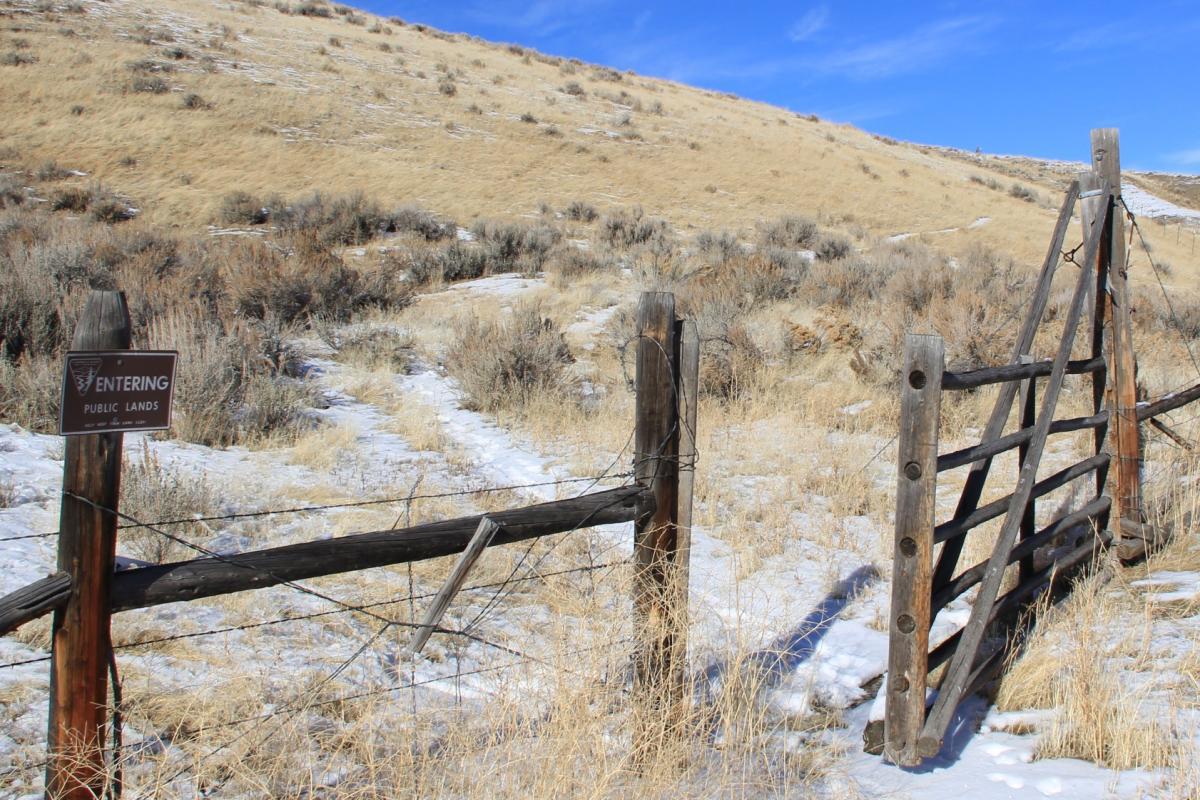 A weathered wooden gate stands open, welcoming visitors to a snowy path leading into a grassy area. A sign on the gate reads "ENTERING PUBLIC LANDS." The landscape is characterized by dry grass and sparse shrubs under a clear blue sky. Cheney Loop mountain bike trail.