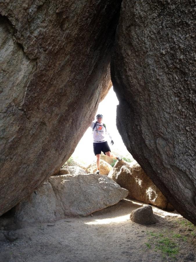 A person standing between two large boulders, with sunlight streaming through the gap. The individual is wearing a helmet and athletic clothing, and they appear to be in a rocky outdoor setting. Pima Road and Dynamite Blvd mountain bike trail.