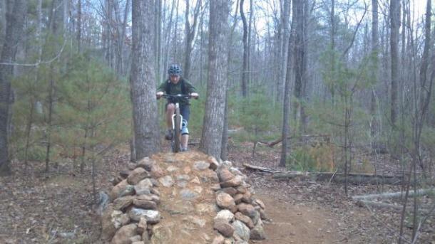 A cyclist wearing a helmet rides over a rocky bridge on a mountain biking trail surrounded by trees. The terrain is natural with a mix of dirt and stones, and the environment is wooded, indicating a forest setting. Woolwine Trails [Shiners Revenge] mountain bike trail.