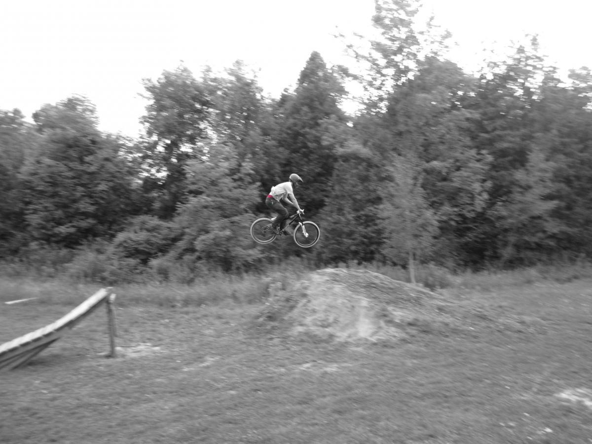 A person wearing a helmet and a t-shirt is mid-air on a bicycle, performing a jump over a dirt ramp in a park surrounded by trees. The image is in black and white. Hanover Freeride Mtb Park mountain bike trail.
