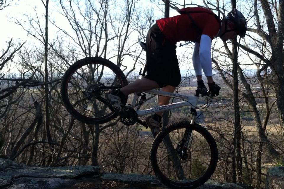 A mountain biker performing a jump on a rocky trail surrounded by bare trees, showcasing an athletic pose as the bike's front wheel is elevated off the ground. The background features a scenic view of the landscape. White Oak Mtn Biology Trails mountain bike trail.
