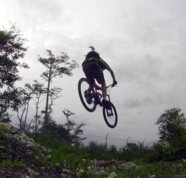 A mountain biker performing a jump on a trail surrounded by trees and cloudy skies. Quiet Waters Park mountain bike trail.