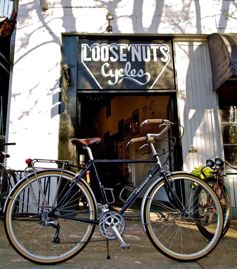 A classic bicycle in front of a shop with a sign reading "Loose Nuts Cycles." The bike features a dark frame, brown leather seat, and retro-style tires. The shop entrance is partially visible, showcasing an inviting interior, with warm lighting and artistic decorations.