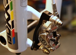 Close-up image of a bicycle brake caliper, showcasing its intricate metal components and connection to the bike frame. The caliper is labeled "XO," indicating its brand, and the colorful design on the frame is partially visible in the background.
