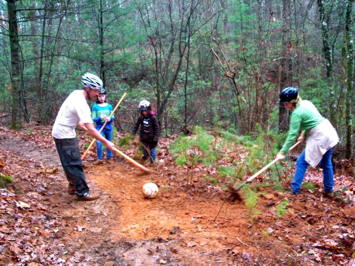 A group of four people, including two children, are working together outdoors to clear a muddy area in a forested environment. They are using tools to remove soil and debris, with a soccer ball visible on the ground. Everyone is wearing helmets for safety. The scene is surrounded by trees and fallen leaves, indicating a rustic setting. Jake to Bull Mountain Connecter mountain bike trail.