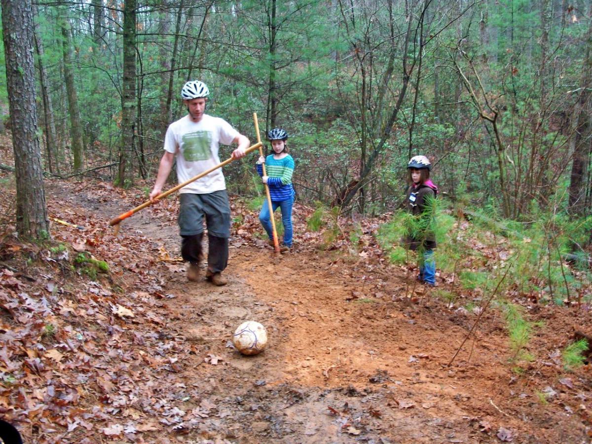 A man and two children are working together to clear a dirt path in a forested area. The man, wearing a helmet and holding a digging tool, is walking along the trail. One child is using a similar tool, while the other stands nearby, observing. The ground is covered with fallen leaves, and a soccer ball is resting on the ground. Young trees and greenery surround the scene. Jake to Bull Mountain Connecter mountain bike trail.