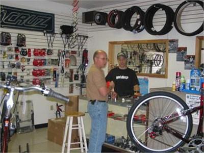 A bicycle shop interior featuring two men interacting over a counter. One man is standing and holding a bicycle part, while the other is behind the counter, looking at him. The shop displays various bicycle components and accessories on the walls, with a focus on wheels and handlebars in the foreground.