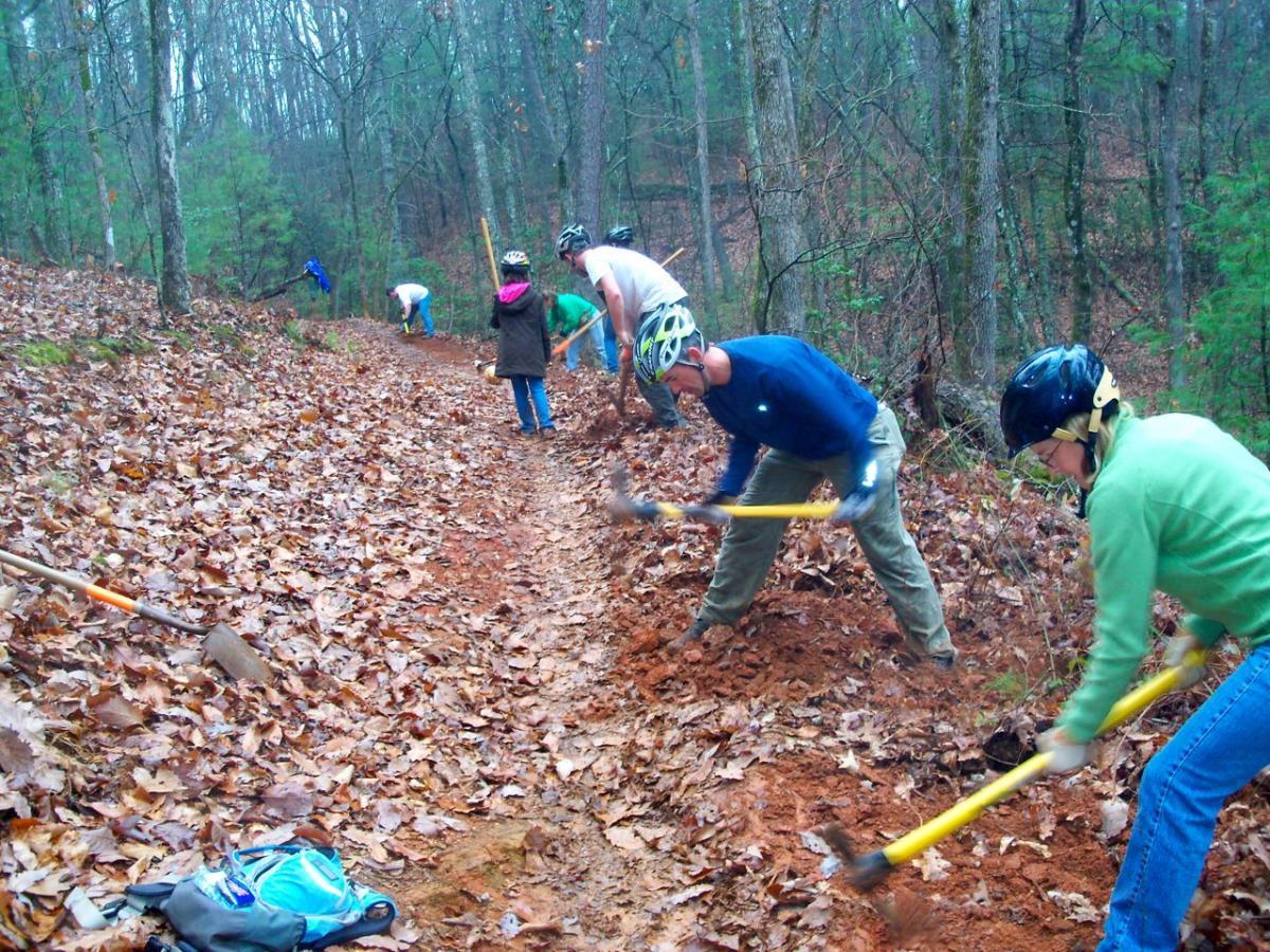 A group of people working collaboratively to improve a dirt trail in a wooded area, using shovels and pickaxes. The scene shows individuals wearing helmets, focused on digging and clearing the path, while surrounded by autumn leaves and trees. Some participants are further down the trail, contributing to the trail maintenance effort. Jake to Bull Mountain Connecter mountain bike trail.