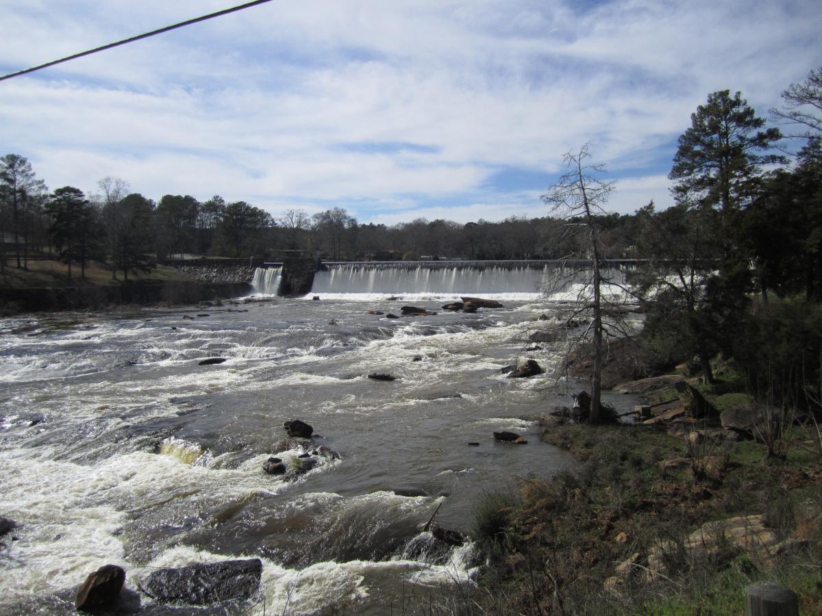 A scenic view of a river with rapids flowing over rocks, leading to a large waterfall in the distance. The shoreline is lined with trees, and a partly cloudy sky can be seen overhead. Dauset Trails Nature Center mountain bike trail.
