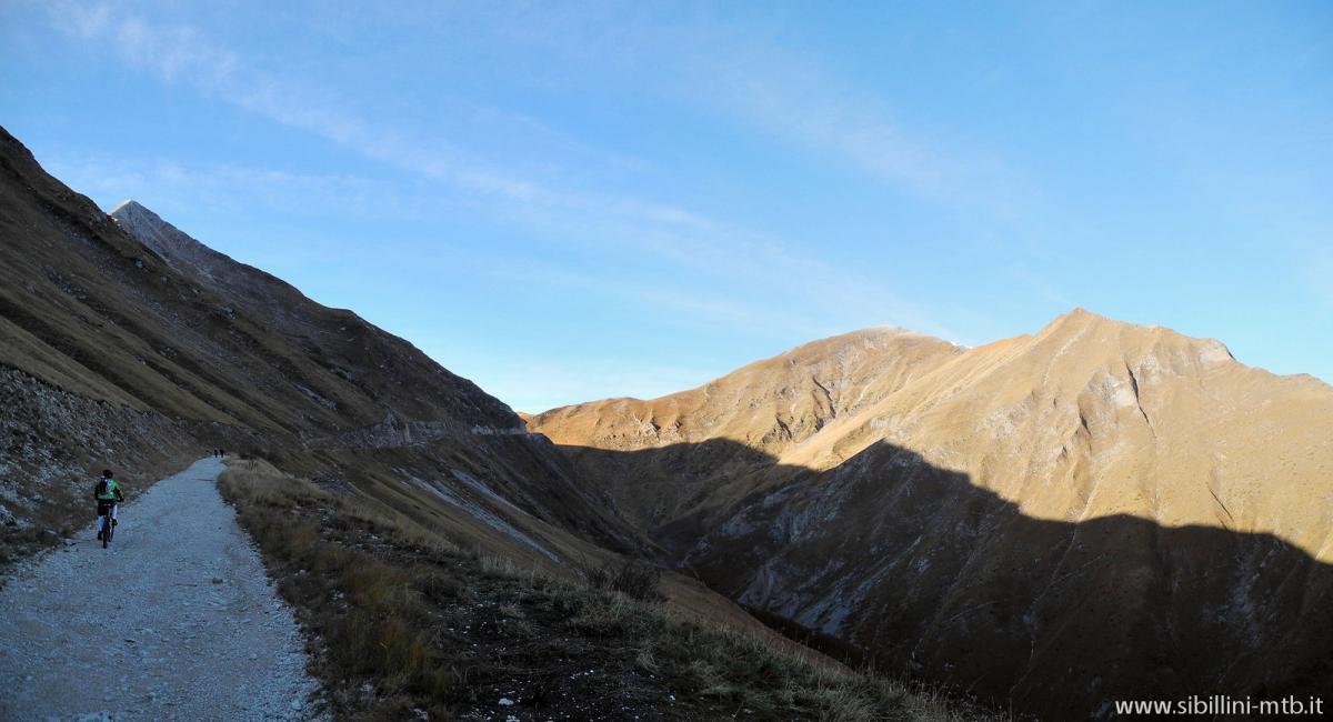 A scenic mountain trail with a cyclist riding along a gravel path. The surrounding landscape features steep, rugged hills and mountains under a clear blue sky, with a mix of sun and shadow highlighting the terrain. Forcella Angagnola mountain bike trail.