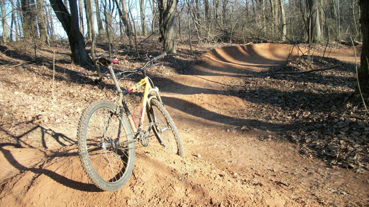 A mountain bike leaning against a dirt jump in a wooded area, with a clear sky and early spring surroundings. brown dirt trails weave through the landscape, lined with bare trees and scattered leaves. Freedom Center mountain bike trail.