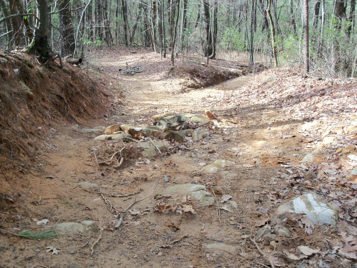 A dirt trail winding through a wooded area, with visible rocks and fallen leaves scattered along the path. A bicycle is lying abandoned on the ground to the left, surrounded by trees with bare branches. Bull / Jake Mountain mountain bike trail.
