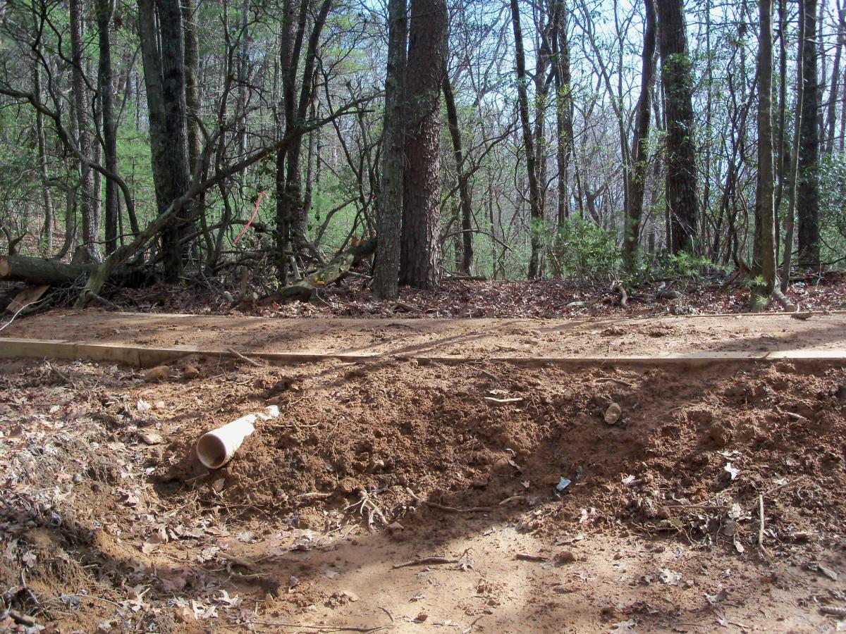 A dirt pathway in a wooded area, showing freshly disturbed soil with a white pipe laying on its side. Surrounding trees are bare of leaves, indicating it is early spring, and some fallen branches and leaves are scattered on the ground. Bull / Jake Mountain mountain bike trail.