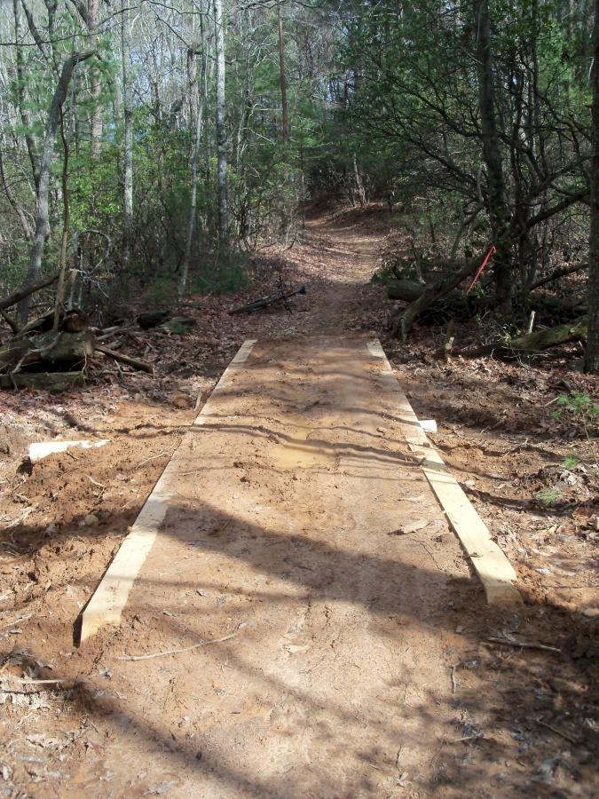 A cleared path through a wooded area featuring a wooden plank pathway. The trail is surrounded by trees and underbrush, with some construction debris visible along the sides. Sunlight filters through the leaves, illuminating the path ahead. Bull / Jake Mountain mountain bike trail.