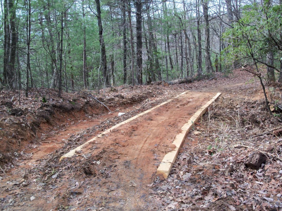 A dirt trail through a forest, featuring wooden planks laid down to create a pathway. Surrounding the trail are trees and scattered leaves on the ground, indicating a natural outdoor setting. Bull / Jake Mountain mountain bike trail.