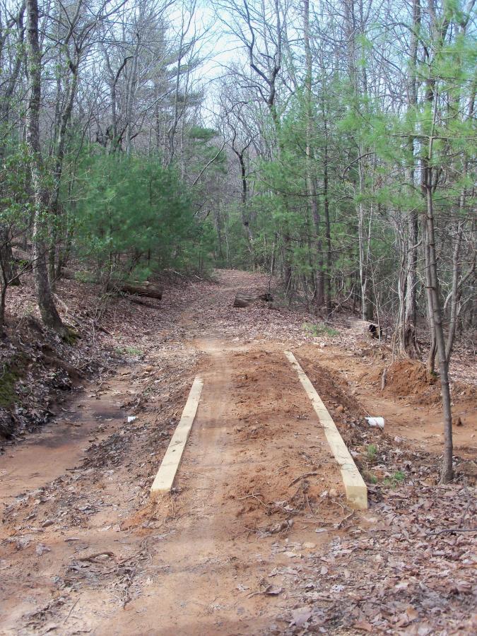 A dirt trail running through a wooded area, flanked by trees and covered in fallen leaves. Two wooden planks are laid across the path, indicating recent construction or maintenance work. Bull / Jake Mountain mountain bike trail.