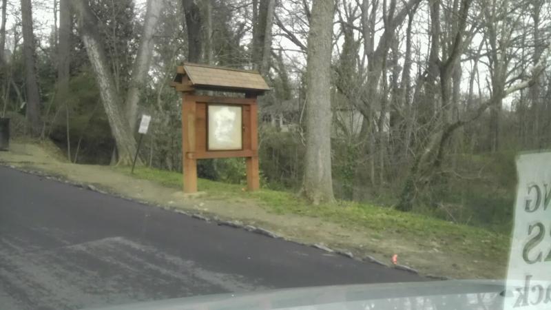 A wooden informational sign with a roof stands beside a road, surrounded by bare trees and greenery. The sign's display area is empty, and there is a glimpse of a creek or stream in the background. The pavement is newly asphalted, showing a fresh layer of blacktop. Morningside Nature Preserve mountain bike trail.