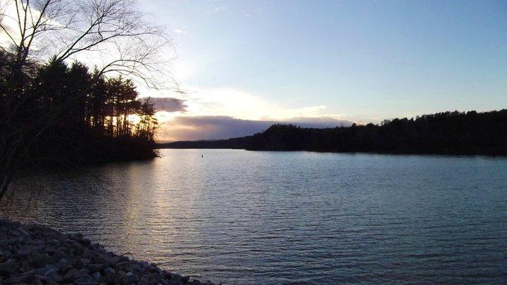 A tranquil view of a lake at sunset, surrounded by trees on the left, with soft ripples in the water reflecting the colors of the sky. The scene captures a peaceful atmosphere, showcasing a blend of blues, purples, and warm tones as the sun sets in the background. Overmountain Victory Trail mountain bike trail.