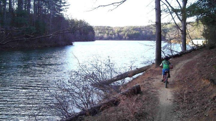 A cyclist riding along a dirt path next to a calm river, surrounded by trees and greenery. The scene captures a tranquil outdoor setting, with the sun shining on the water and a peaceful landscape in the background. Overmountain Victory Trail mountain bike trail.