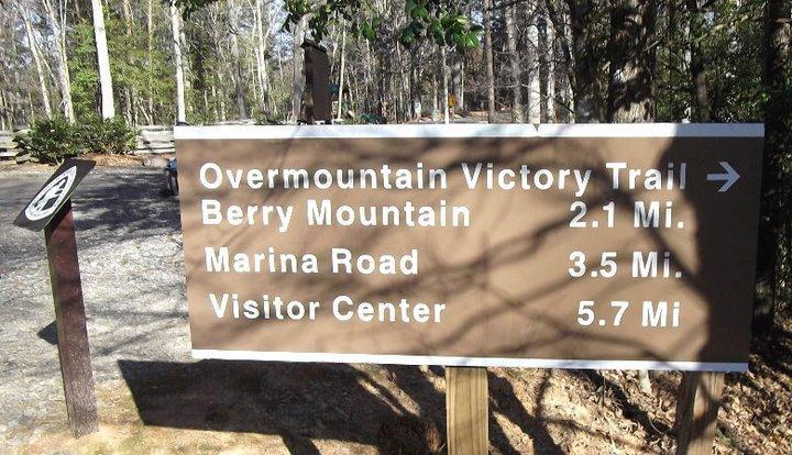 Brown directional sign showing distances to various locations: Overmountain Victory Trail, 2.1 miles; Berry Mountain, 3.5 miles; Marina Road, and Visitor Center, 5.7 miles. Trees and a gravel path are visible in the background. Overmountain Victory Trail mountain bike trail.