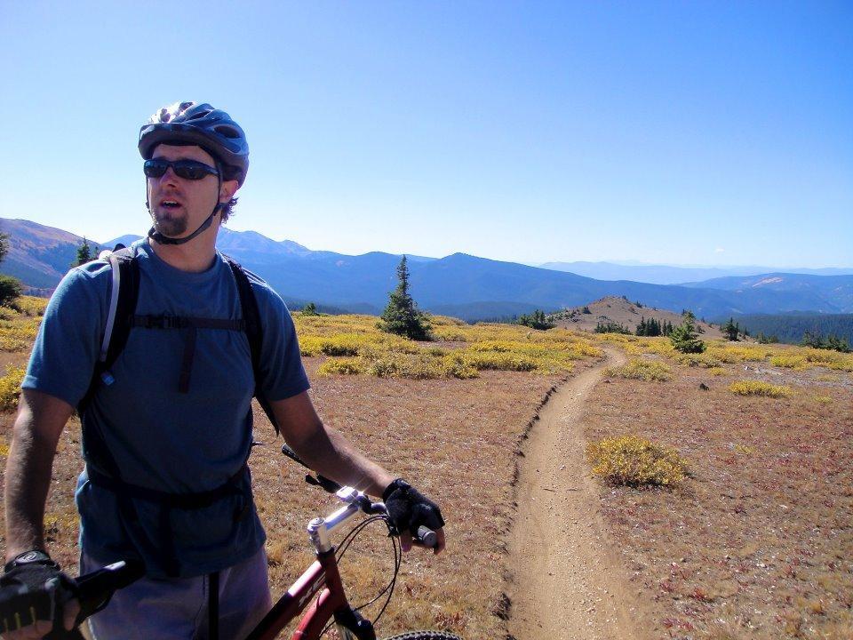 A mountain biker wearing a helmet and sunglasses stands beside their bike on a trail surrounded by autumn-colored foliage and scenic mountain views under a clear blue sky. Monarch Crest Trail mountain bike trail.
