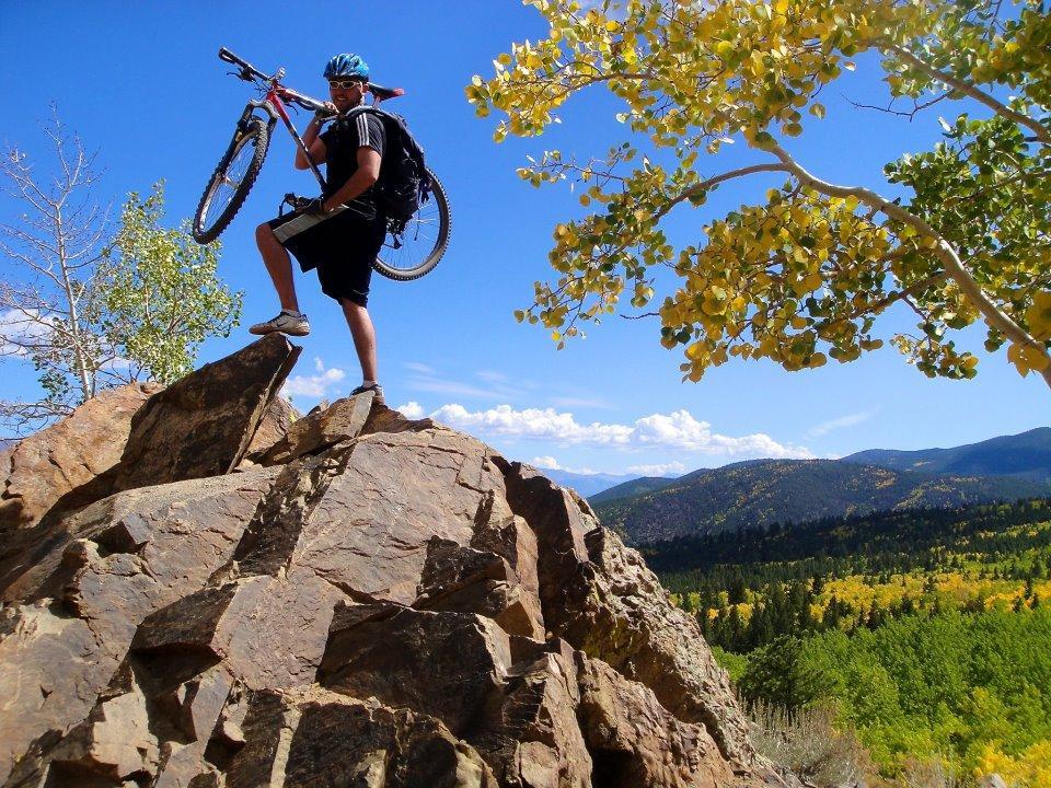 A person stands atop a large rock, holding a mountain bike above their head. The individual wears a blue helmet and a backpack, and is dressed in athletic attire. The scene features a vibrant blue sky with fluffy white clouds and expansive mountain views in the background, adorned with trees displaying autumn colors. Monarch Crest Trail mountain bike trail.