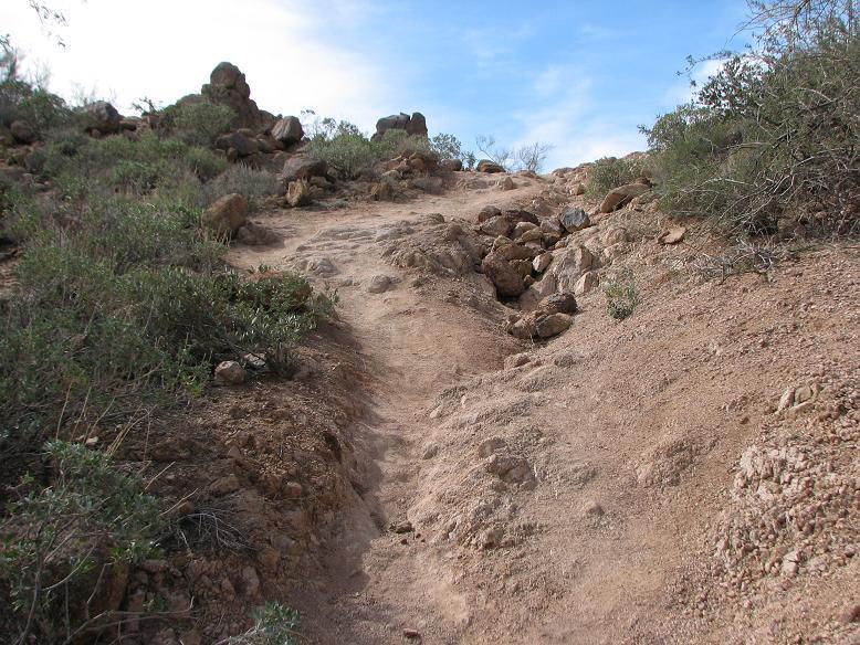A rugged dirt path leading up a hillside, surrounded by rocky terrain and sparse vegetation under a partly cloudy sky. Cat Peaks Via Ruidoso mountain bike trail.