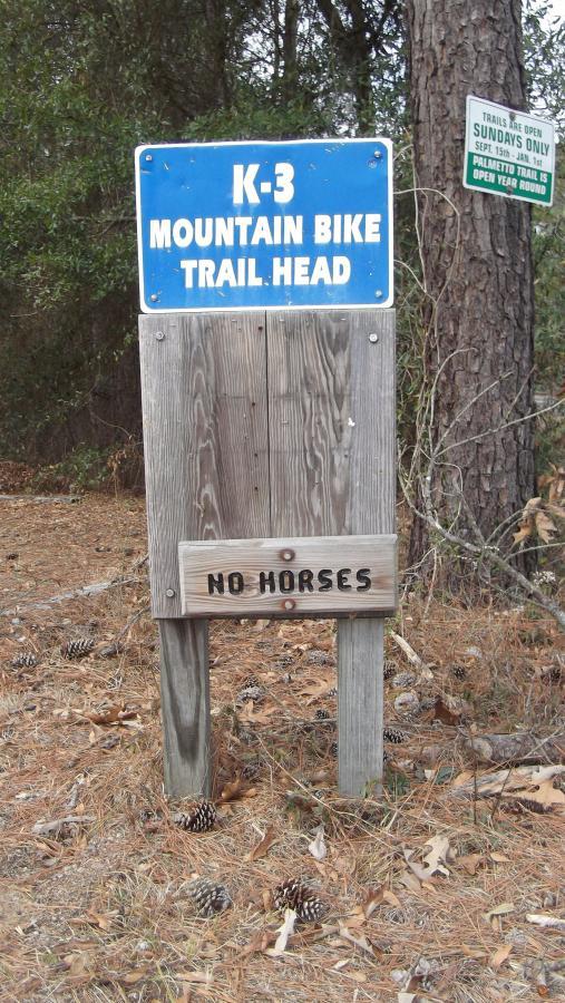 Signpost marking the K-3 Mountain Bike Trail Head, with a blue background indicating trail information. Below, a wooden plank states "No Horses." Surrounding area features pine needles and cones, with trees in the background. Killer Three Loop mountain bike trail.