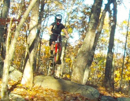 A mountain biker in a black helmet and gear jumps off a large rock in a forested area filled with autumn foliage. Vietnam Trails mountain bike trail.