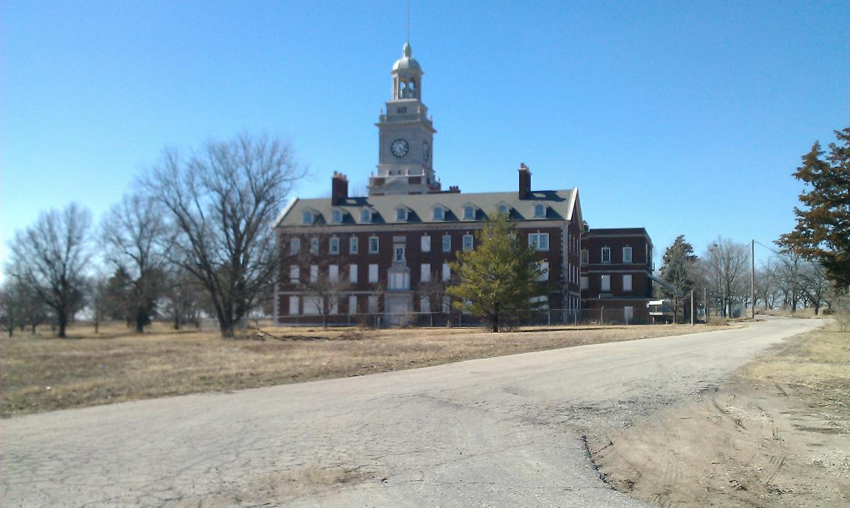 An old brick building with a clock tower, surrounded by trees and a grassy area. A gravel road leads up to the building, which appears vacant. The sky is clear and blue, hinting at a sunny day. Maclennan Park mountain bike trail.