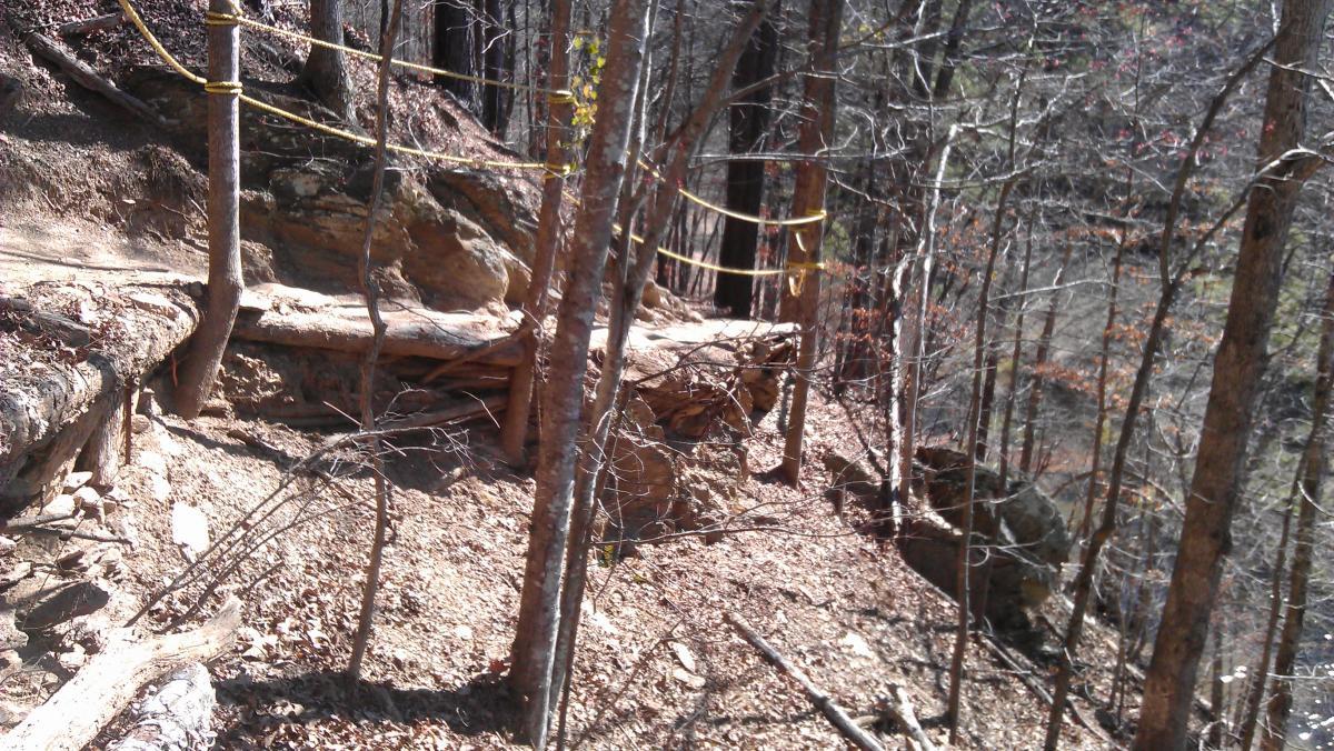 A winding trail through a wooded area, with exposed rocky terrain and tree trunks. A log is visible along the path, and yellow caution tape can be seen tied around nearby trees, indicating a potentially hazardous or restricted section of the trail. The ground is covered in fallen leaves and loose dirt. Van Michael Trail mountain bike trail.