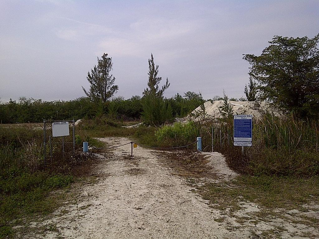 A sandy path leading through a fenced area with a sign indicating an entrance. Lush greenery and trees are visible on both sides, while a small hill of sand or dirt is seen in the background. There are informational signs on either side of the path, and the sky appears overcast. West Delray Regional Park mountain bike trail.
