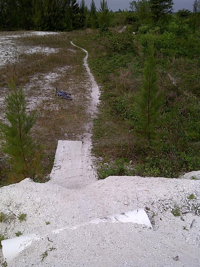 A dirt bike ramp made of concrete is positioned at the edge of a slope, leading down into a grassy area. A narrow, winding path can be seen on the left side, surrounded by sparse vegetation and small trees. In the background, more greenery is visible under a cloudy sky. There is a blue bicycle lying on the ground near the ramp. West Delray Regional Park mountain bike trail.
