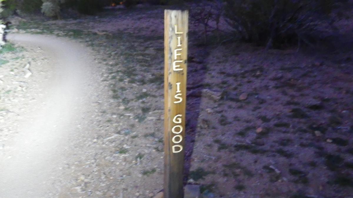 Wooden signpost along a dirt path with the engraved message "LIFE IS GOOD." The surrounding area features sparse vegetation and a natural, earthy landscape. Fantasy Island mountain bike trail.